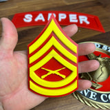 USMC enlisted rank insignia in hand, featuring Gunnery Sergeant rank in red and yellow design with crossed rifles.