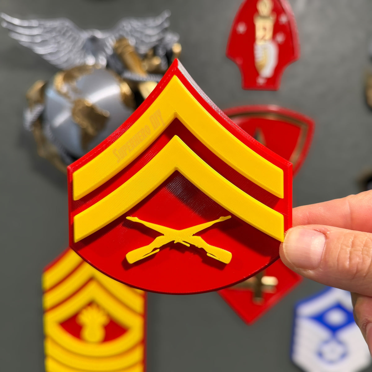 Hand holding a USMC Corporal rank insignia against a dark gray wall, other insignia displays blurred behind